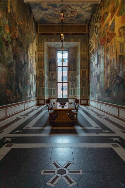 The interior of Oslo City Hall features intricate murals on the walls surrounding a central wooden table. Natural light streams in through large windows, highlighting the artistic details.