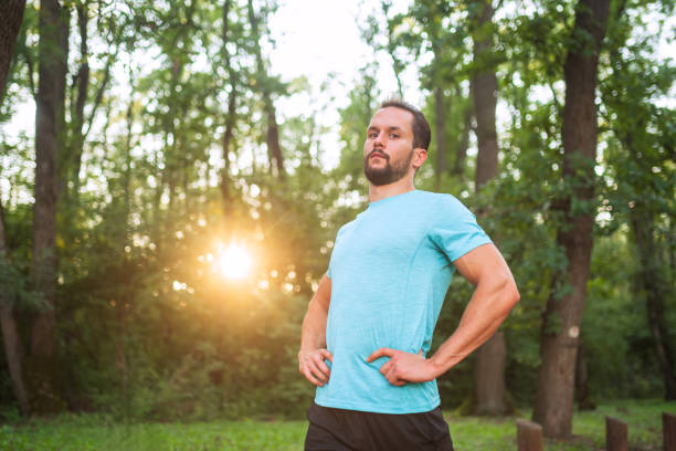 Young sportsman is posing with hands on hips in a forest at sunset, he is wearing a blue t-shirt. He seems satisfied after a workout session