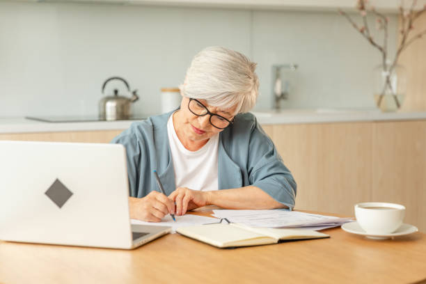 istock Middle aged senior woman using laptop computer writing notes at home. Focused mature old businesswoman work in home office making written records doing online work. Mature boss professional worker. 2179954203