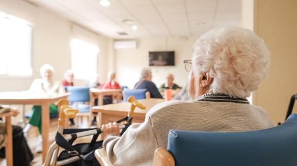 Elderly woman with foldable walker in nursing home Back view of senior woman with short white hair sitting on chair with foldable walker with friends in blurred background at nursing home nursing home stock pictures, royalty-free photos & images