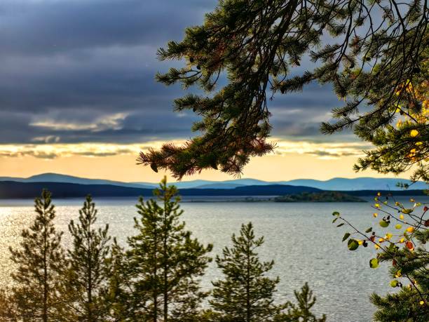 The tranquil lake reflects warm sunset colors, framed by lush evergreen trees and rolling mountains in the distance, creating a serene landscape.