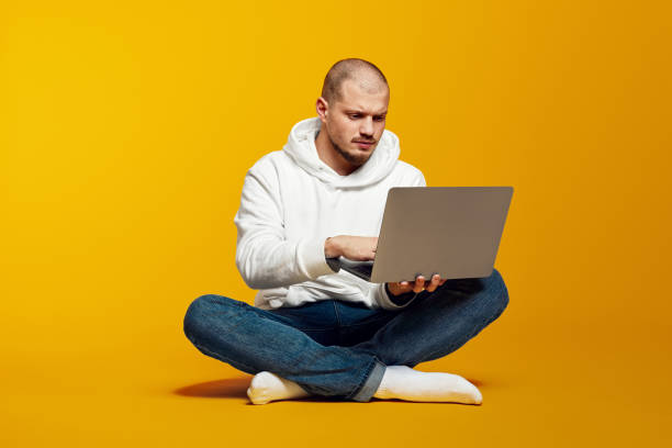 istock Young serious caucasian man sitting on floor and using modern notebook isolated on yellow background 2179999661
