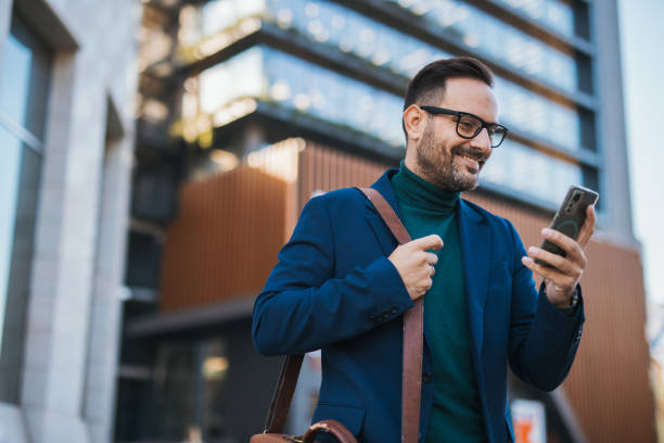 istock A handsome, well-dressed business man stands in front of a modern building, using his phone to write a business email. It’s morning, and he is engaged in work obligations while enjoying the beautiful day and feeling positive. 2180001991