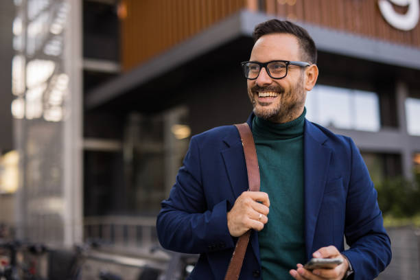 istock A handsome, well-dressed business man stands in front of a modern building, using his phone to write a business email. It’s morning, and he is engaged in work obligations while enjoying the beautiful day and feeling positive. 2180009521