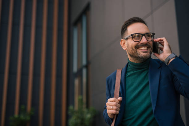 istock handsome, well-dressed business man stands in front of a modern building, using his phone to write a business email. It’s morning, and he is engaged in work obligations while enjoying the beautiful day and feeling positive 2180020700