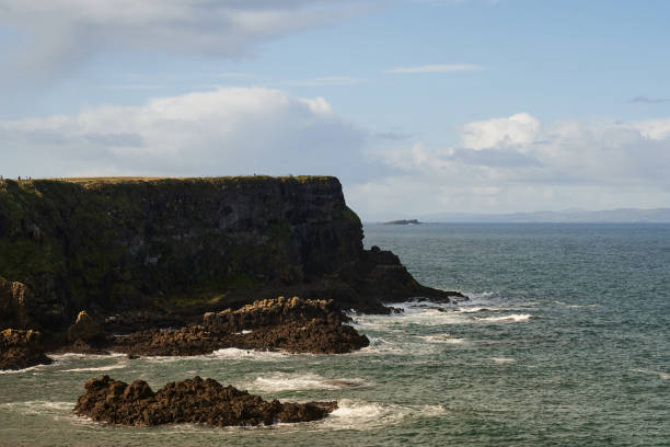 A serene view of the Irish coastline during the morning hours. The rugged cliffs stand tall above the rocky shore, as gentle waves crash against the coast. The morning light enhances the natural beauty of the scene, creating a peaceful atmosphere. The distant horizon, rolling ocean, and rocky textures of the shore offer a glimpse of the untamed landscape of Ireland's coastal regions, making this image a perfect representation of tranquility and natural beauty.