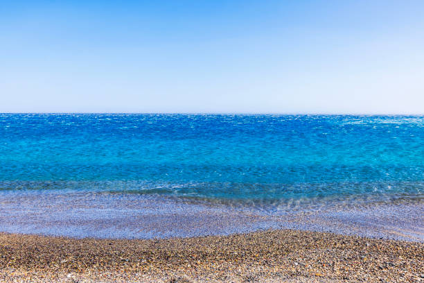 Clear blue Mediterranean sea with small pebbles on shore under sunny sky, with calm water stretching to horizon.