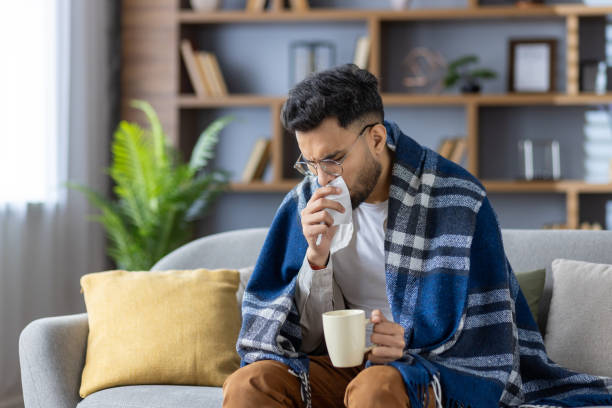 young man sitting on sofa covered with blanket, drinking hot beverage and blowing nose, feeling unwell and cold - grip stok fotoğraflar ve resimler