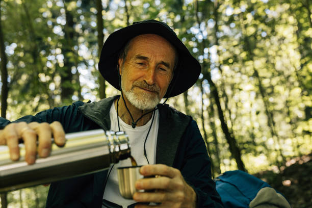 istock Senior male in hat pouring coffee from a thermos while taking a break in a forest during walk 2180046118