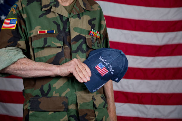 American veteran holding cap over heart wearing  army camouflage jacket with award ribbons, American Flag in background.