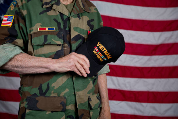 American veteran holding cap over heart wearing  army camouflage jacket with award ribbons, American Flag in background.