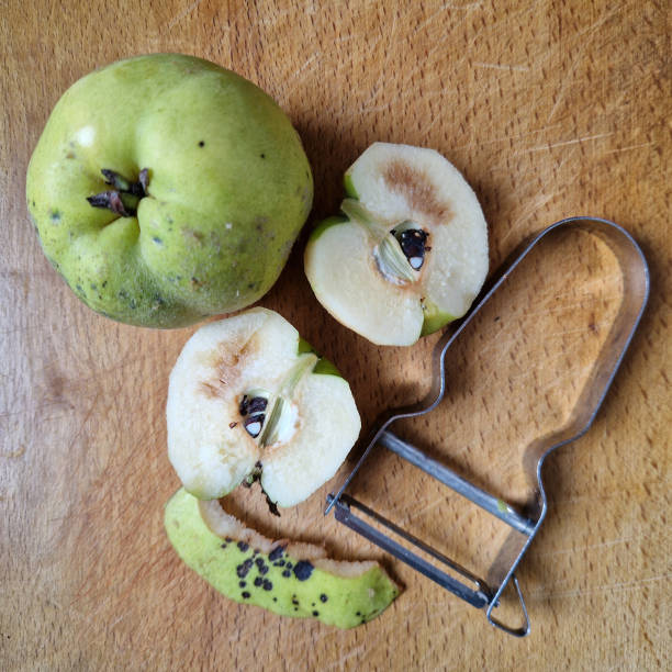 A close-up image of a quince fruit on a wooden cutting board, with one half sliced open to reveal the seeds inside. A vegetable peeler lies next to the peeled skin, emphasizing the process of preparing fresh quince for cooking or baking. The rustic feel and natural textures make this image perfect for themes related to home cooking, organic produce, seasonal fruits, and kitchen preparation.