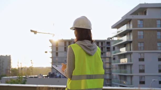 istock Female construction engineer with a hard hat and safety vest is writing on a clipboard while inspecting a construction site in the evening at sunset 2180060931