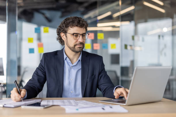 istock Mature financier reviewing paperwork while using laptop in modern office setting with focus on investment strategy and financial analysis 2180067997