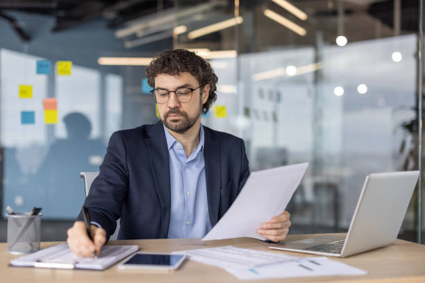 istock Mature financier working diligently inside modern office reviewing paperwork with focus and precision using laptop and smartphone 2180068406