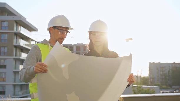 istock Two architects wearing safety hard hats are holding blueprint and discussing something on construction site at sunrise. Architect and teamwork concept 2180070793