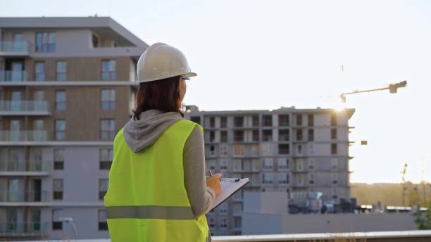istock Female construction engineer with a hard hat and safety vest is writing on a clipboard while inspecting a construction site in the evening at sunset 2180078683