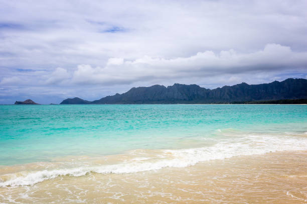 Soft waves lap against the shore, with vibrant turquoise waters and a backdrop of mountains under a moody sky.