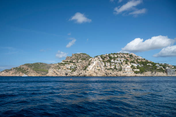 A beautiful view of Mallorca's hillside residences perched on rocky cliffs, overlooking the calm blue Mediterranean Sea. The lush greenery and bright sky enhance the scenic landscape, a picturesque example of coastal living in the Balearic Islands.