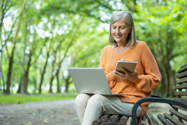 istock Senior woman smiling while using a laptop and notebook in a park, enjoying technology and writing outdoors 2180082979
