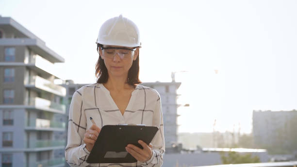 istock Woman construction engineer wearing white checked blouse and hard hat is making notes on a clipboard while inspecting a building site at sunset, front view. Architecture and engineering concepts 2180092934