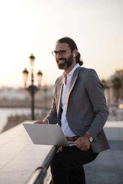 Businessman using laptop outdoors by the sea is smiling at sunset