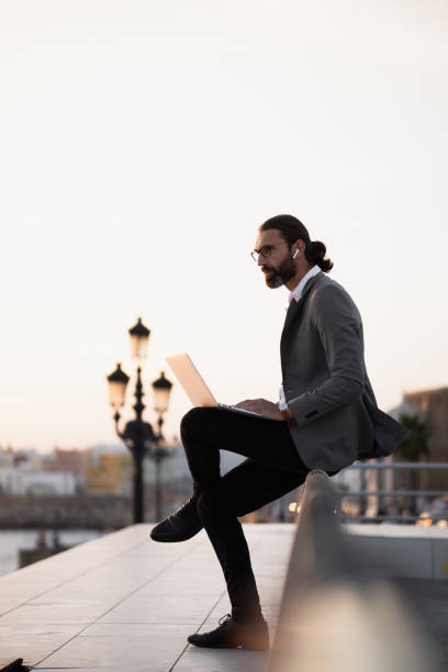 istock Businessman working on laptop outdoors sitting on concrete ledge at sunset 2180104779