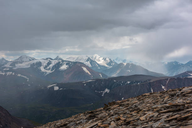 istock Dramatic top view from stone pass above alpine valley to large mountain range with snow-capped peaks on horizon in rain in gray cloudy sky. Rocky steep slope and snowy tops far away under rainy clouds 2180109779