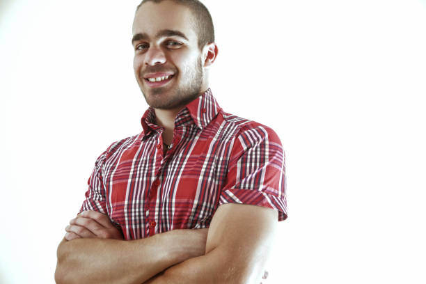 Confident, relaxed young man in a red shirt smiles with crossed arms against a white background, exuding happiness and contentment