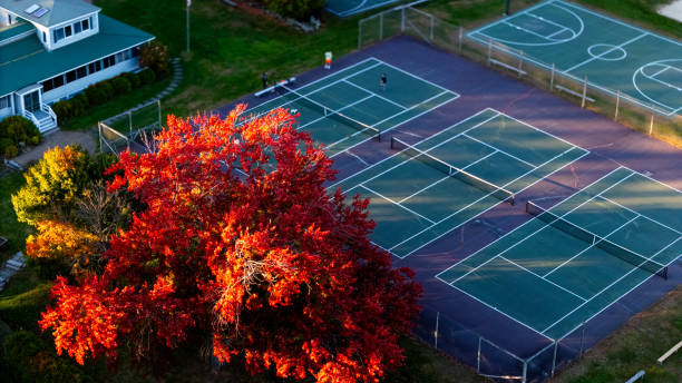 Aerial view of tennis courts surrounded by vibrant autumn trees next to a peaceful lakeside. The scene captures the golden and red hues of fall, with a serene body of water in the background and a basketball court in the foreground, creating a tranquil outdoor sports setting. It's the best photo of the year.