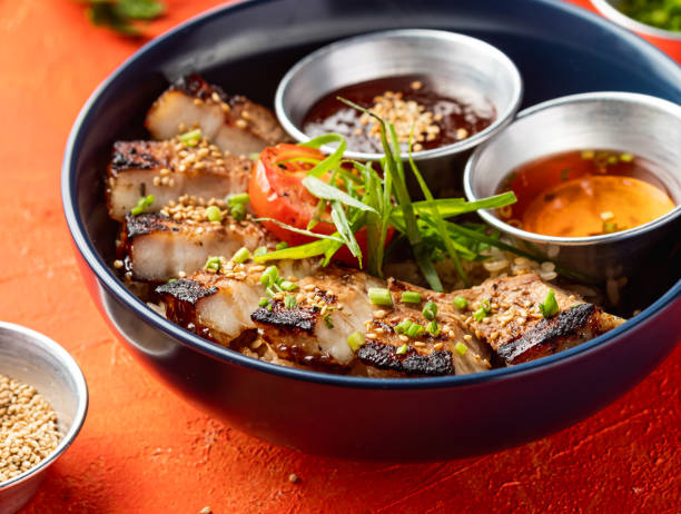 Korean Grilled Pork Belly Rice Bowl with dipping sauce, Coriander and sesame seeds isolated on orange background closeup side view of fast food meal