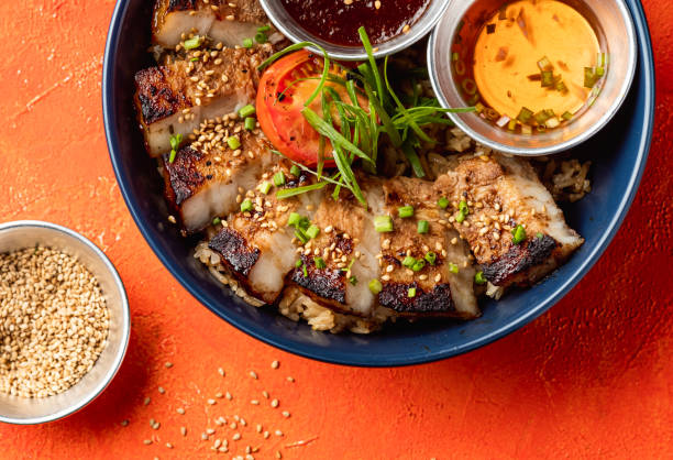 Korean Grilled Pork Belly Rice Bowl with dipping sauce, Coriander and sesame seeds isolated on orange background closeup top view of fast food meal