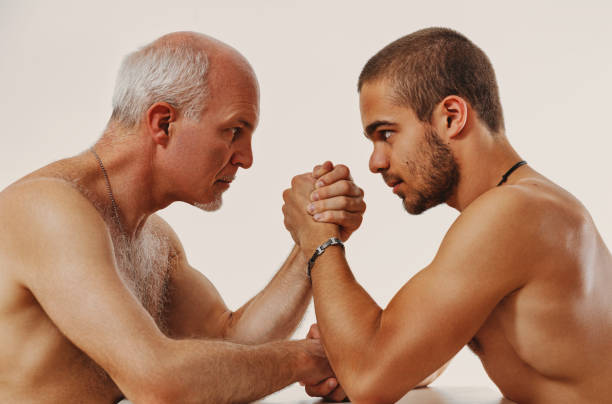 Two shirtless men fiercely arm wrestle, showing intense effort and determination. The close up shot captures their masculinity and physicality in a tense struggle