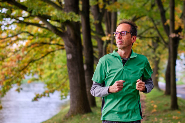 A man jogs along a scenic riverside path, embracing the tranquility of nature. Dressed in a emerald green t shirt, he enjoys the crisp morning air, exuding a sense of calm and vitality. The lush, green trees overhead create a serene backdrop, highlighting his commitment to self-care and fitness.