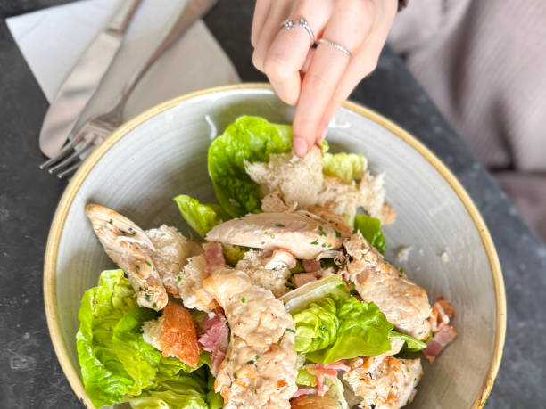 Close-up image of unrecognisable young woman eating Chicken Caesar salad, crispy green Romaine lettuce leaves with chopped bacon pieces, sliced chicken breast, croutons (toasted white bread pieces), black marble table top background, elevated view