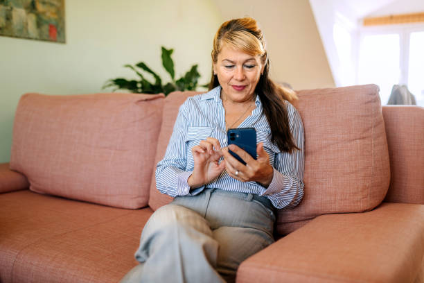 istock Casually clothed mature woman using smart phone while sitting on a sofa at home 2180197851