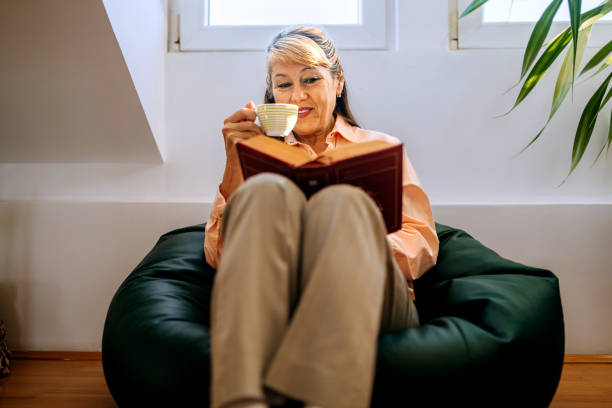 istock Well dressed senior woman drinking coffee and reading book while relaxing on a bean bag at home 2180198785