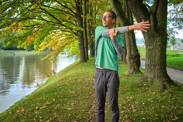 A man stands beside a tree, stretching his arms and focusing on his breathing, immersed in the peacefulness of the natural setting. The vibrant autumn foliage and calm river in the background reflect his mindful approach to self-care. The image captures a quiet, focused moment of health and relaxation, symbolizing balance and rejuvenation in nature