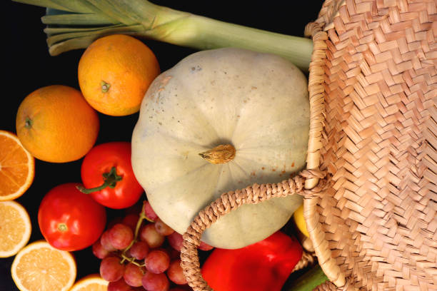 Round straw bag and various seasonal fruits and vegetables on dark background. Summer and fall produce. Top view.