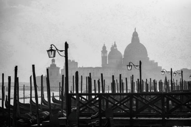 This mid-shot captures a series of weathered wooden mooring posts and an antique lamppost in the foreground, bordering a Venetian canal. In the hazy background, the dome of a distant basilica is faintly visible, enveloped in atmospheric fog.
