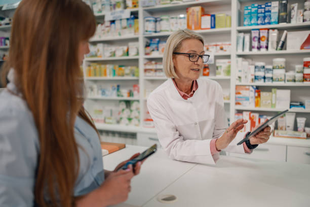 istock Senior pharmacist at chemist's shop with tablet serving customer. 2180214008
