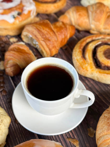 istock Full frame image white, china coffee cup and saucer full of black coffee, assorted, breakfast pastries on brown wood grain surface, iced Belgian buns topped with glacé cherry, cinnamon swirl pastries, scones and all butter croissants, roasted coffee beans 2180217769