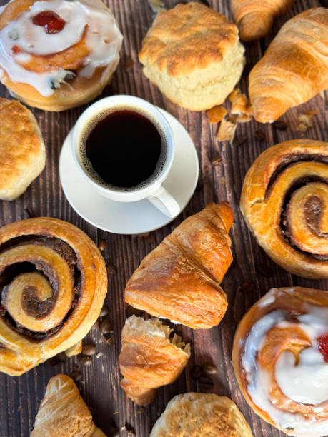 istock Full frame image white, china coffee cup and saucer full of black coffee, assorted, breakfast pastries on brown wood grain surface, iced Belgian buns topped with glacé cherry, cinnamon swirl pastries, scones and all butter croissants, roasted coffee beans 2180217774
