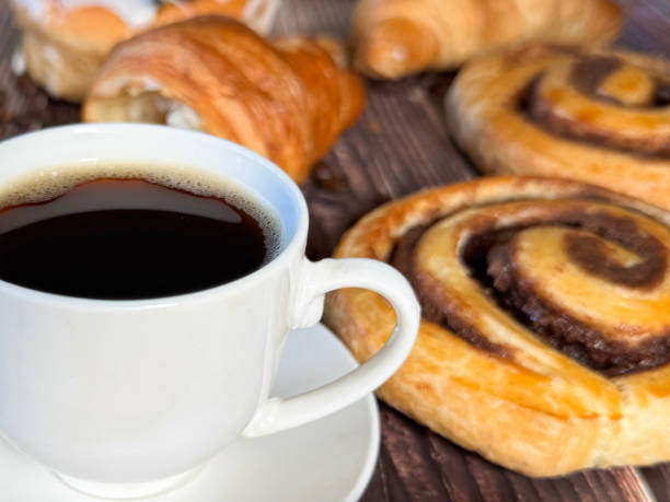 istock Full frame image white, china coffee cup and saucer full of black coffee, assorted, breakfast pastries on brown wood grain surface, iced Belgian buns topped with glacé cherry, cinnamon swirl pastries, scones and all butter croissants, roasted coffee beans 2180217775