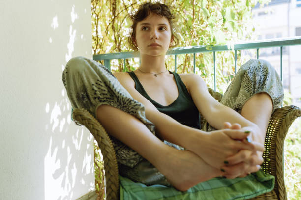 Portrait meditating young woman, teen girl, closed eyes, brown curly hair tied in ponytail, in wide trousers with Mandala pattern, in loose clothing, sitting in wicker chair on terrace, enjoying sun