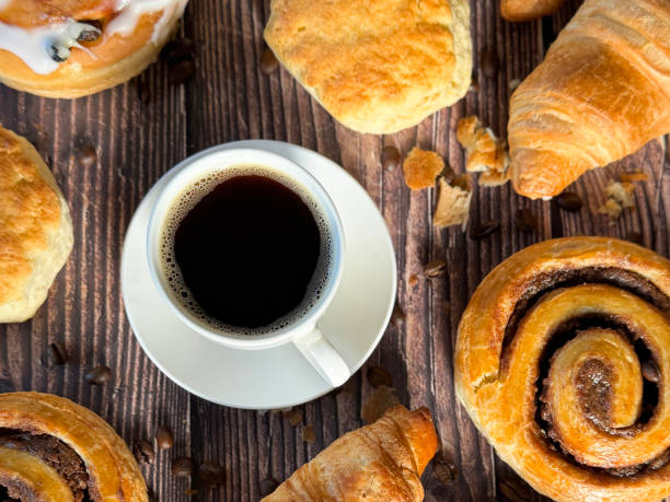Stock photo showing close-up, elevated view of a white, china coffee cup and saucer of Americano coffee (espresso and water) surrounded by freshly baked, iced Belgian buns, cinnamon swirl pastries, scones, all butter croissants and roasted coffee beans, on brown wood grain background, ready to be eaten for breakfast.