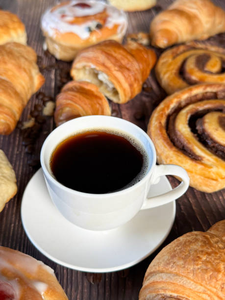 istock Full frame image white, china coffee cup and saucer full of black coffee, assorted, breakfast pastries on brown wood grain surface, iced Belgian buns topped with glacé cherry, cinnamon swirl pastries, scones and all butter croissants, roasted coffee beans 2180218205