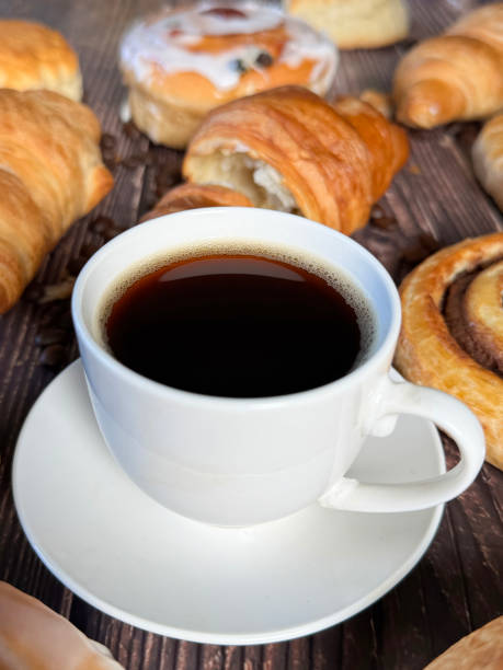 istock Full frame image white, china coffee cup and saucer full of black coffee, assorted, breakfast pastries on brown wood grain surface, iced Belgian buns topped with glacé cherry, cinnamon swirl pastries, scones and all butter croissants, roasted coffee beans 2180218207