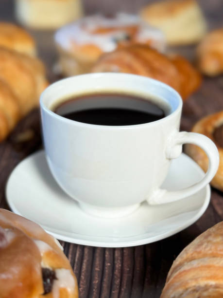 istock Full frame image white, china coffee cup and saucer full of black coffee, assorted, breakfast pastries on brown wood grain surface, iced Belgian buns topped with glacé cherry, cinnamon swirl pastries, scones and all butter croissants, roasted coffee beans 2180218208