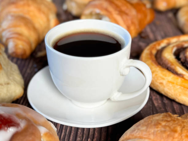 istock Full frame image white, china coffee cup and saucer full of black coffee, assorted, breakfast pastries on brown wood grain surface, iced Belgian buns topped with glacé cherry, cinnamon swirl pastries, scones and all butter croissants, roasted coffee beans 2180218212